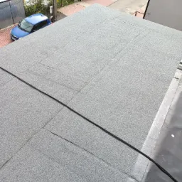 Szary dach pokryty papą termozgrzewalną, widok z góry na płaską powierzchnię z widocznymi łączeniami papy i fragmentem rolki papy w rogu, w tle błękitny samochód i zabudowa jednorodzinna.