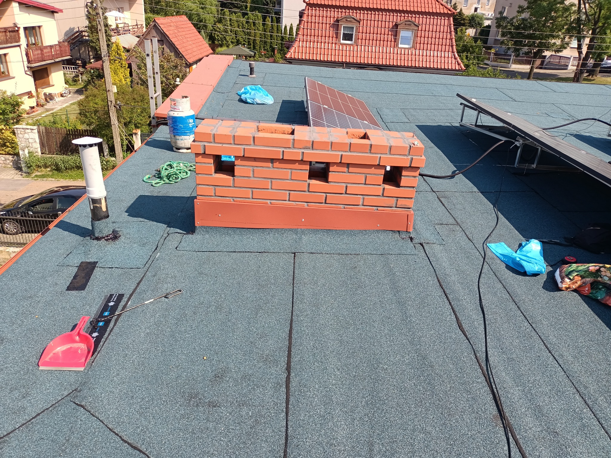 Nowy, płaski dach pokryty papą z widocznym kominem z cegły, panelami słonecznymi, butlą gazową i narzędziami dekarskimi.