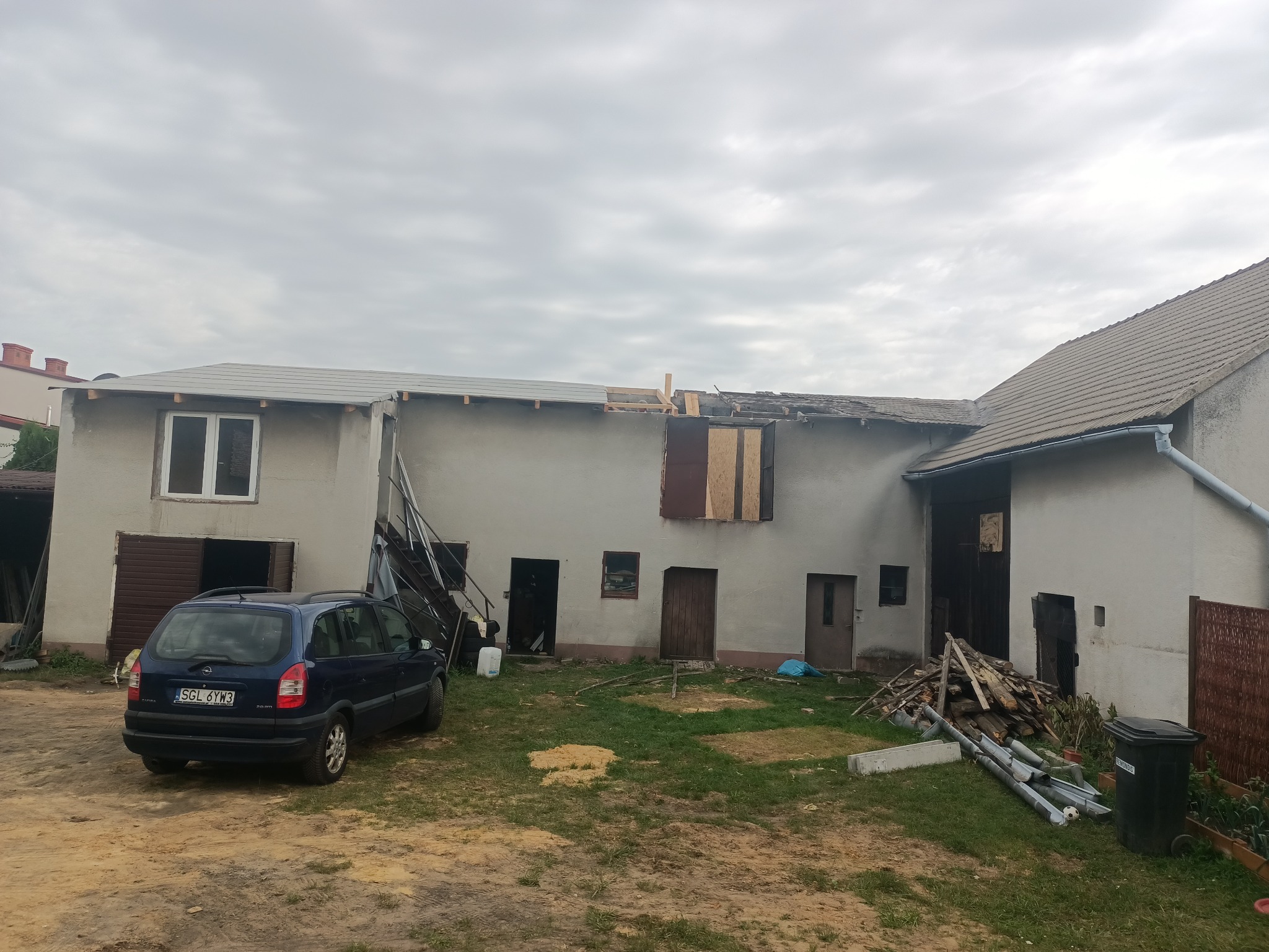 Dom w trakcie wymiany dachu: częściowo zdjęte stare pokrycie, widoczne nowe fragmenty dachu, niebieski samochód osobowy na podwórku.