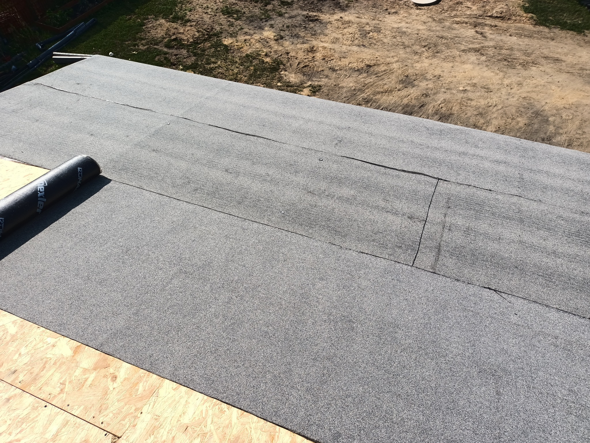 Fragment dachu pokrytego papą termozgrzewalną z widocznym zakładem i rolką materiału na drewnianym podłożu OSB.