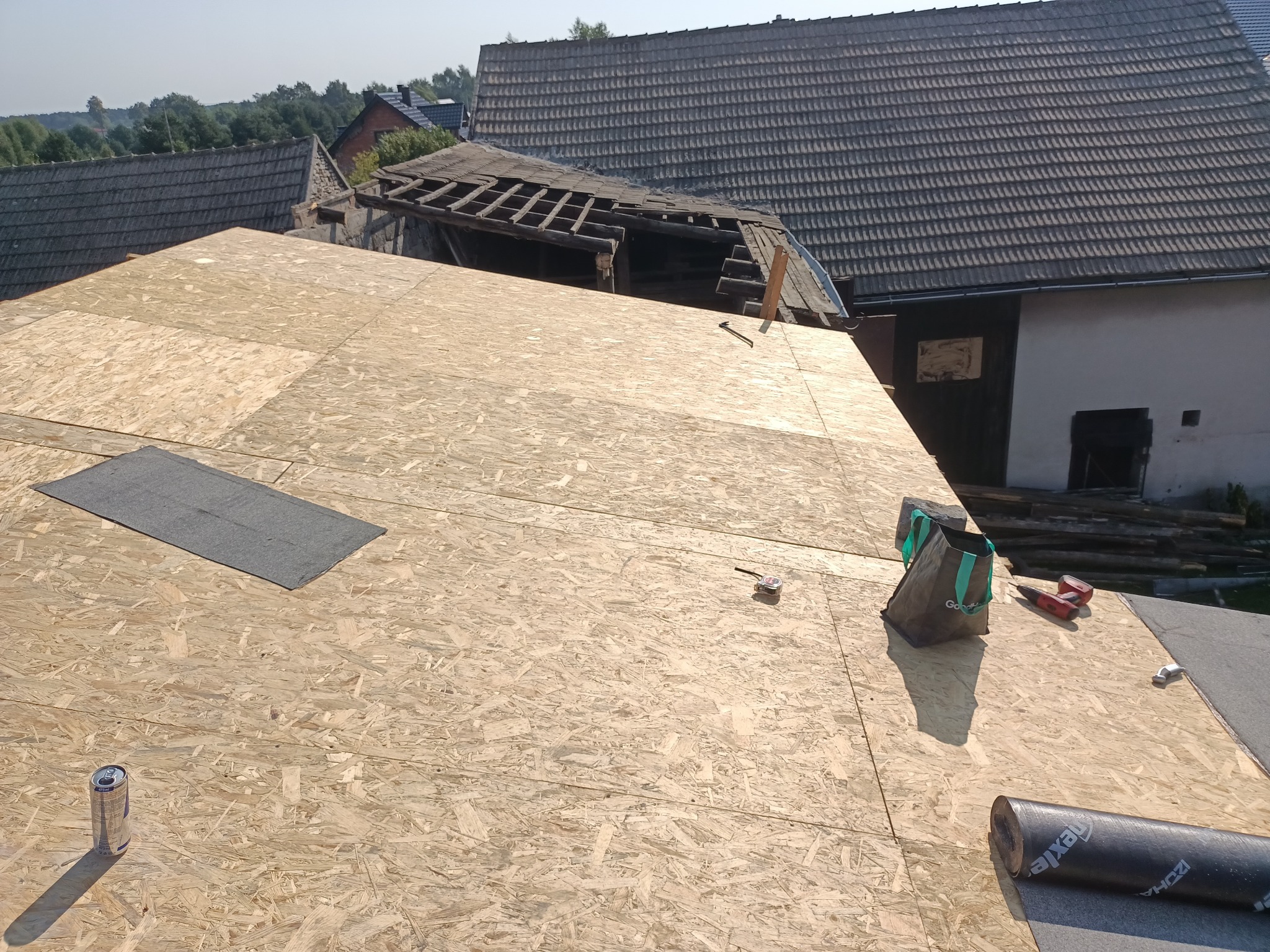 Przygotowanie do pokrycia dachu: płyty OSB na konstrukcji, widoczne narzędzia, rolka papy i częściowo rozebrany stary dach w tle.