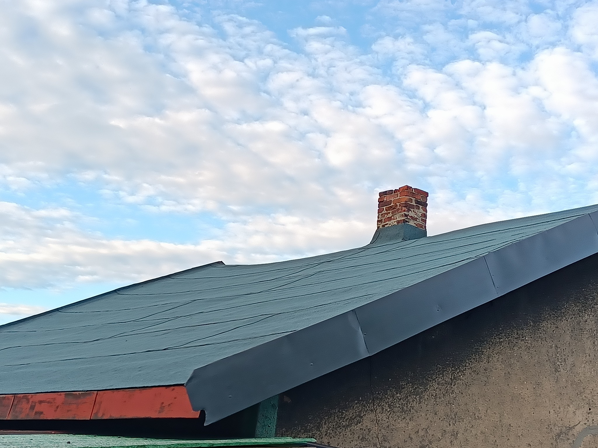 Dach pokryty szarym materiałem bitumicznym, widoczny ceglany komin na tle błękitnego, pochmurnego nieba, okap z obróbką blacharską.