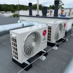 Dwa białe, zewnętrzne moduły klimatyzacji LG Dual Inverter, zamontowane na płaskim dachu z papy, z widocznymi przewodami wentylacyjnymi i kominami w tle.