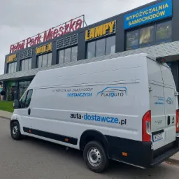 Biały samoch&oacute;d dostawczy z logo PlazaAuto i adresem auta-dostawcze.pl, zaparkowany przed budynkiem Retail Park Mieszka z widoczną reklamą wypożyczalni samochod&oacute;w.