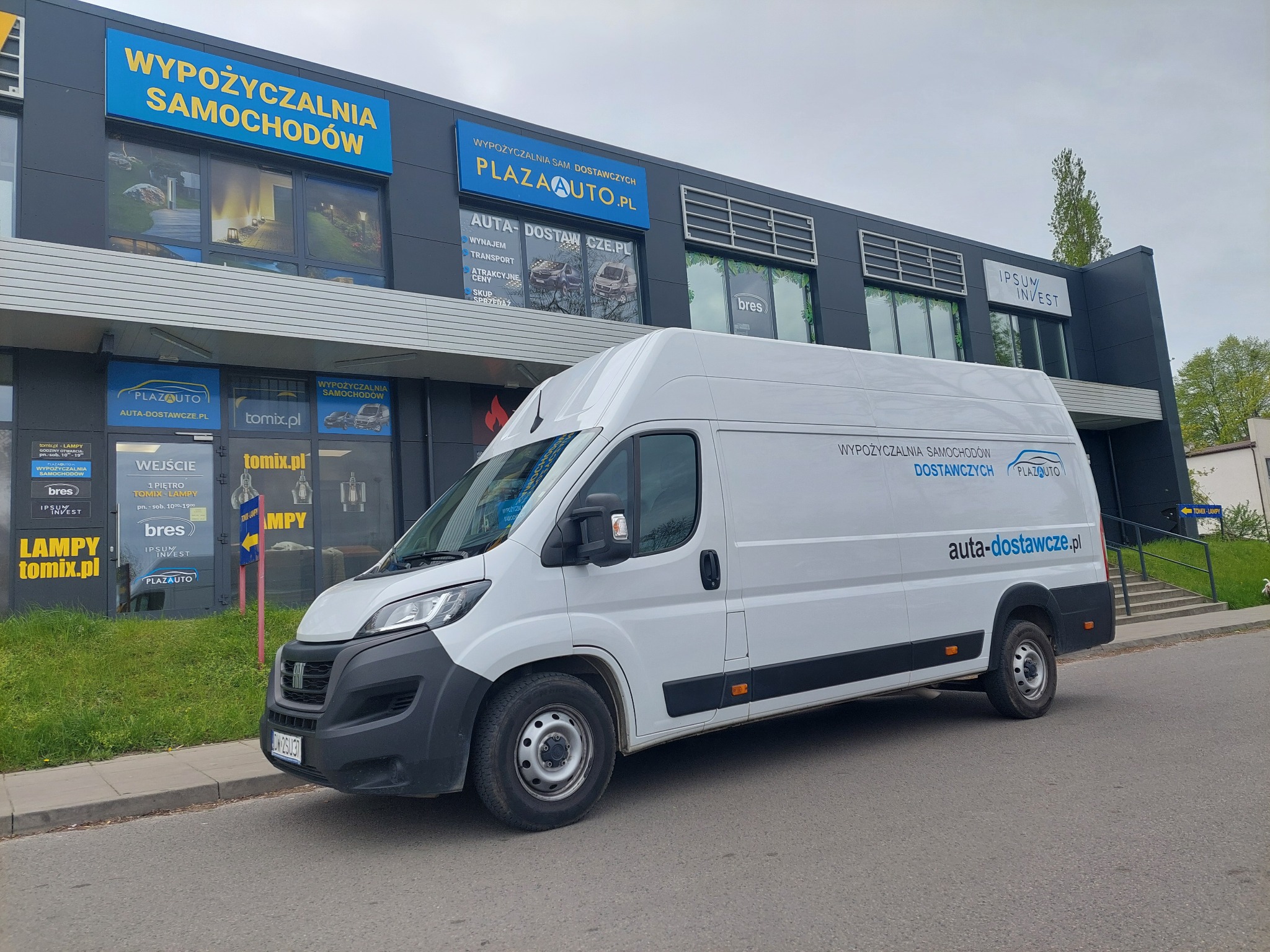 Biały samochód dostawczy zaparkowany przed budynkiem z szyldami firm, w tym z napisem 'WYPOŻYCZALNIA SAMOCHODÓW DOSTAWCZYCH' i adresem strony auta-dostawcze.pl.