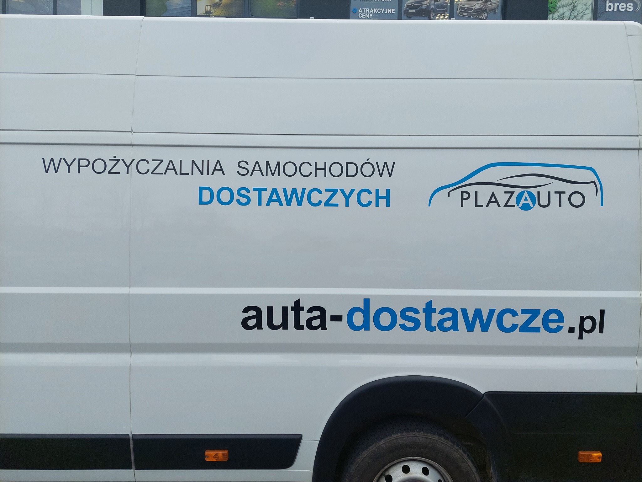 Biały samochód dostawczy z reklamą firmy PlazaAuto, oferującej wynajem aut dostawczych, z widocznym adresem strony internetowej.