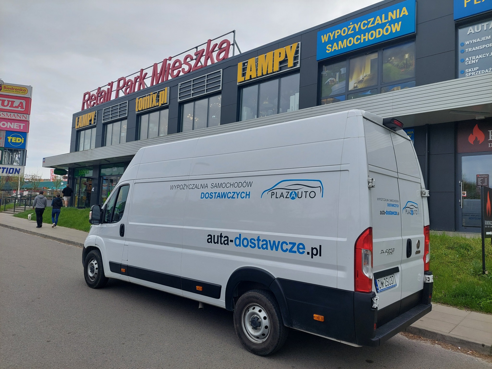 Biały samochód dostawczy z logo PlazaAuto i adresem auta-dostawcze.pl, zaparkowany przed budynkiem Retail Park Mieszka z widoczną reklamą wypożyczalni samochodów.