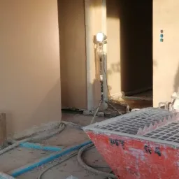 Wnętrze w trakcie prac wykończeniowych: świeżo otynkowane ściany w odcieniach beżu, widoczny agregat tynkarski z resztkami zaprawy, przewody oraz oświetlenie halogenowe na statywie.