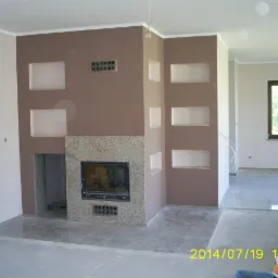 Wnętrze pokoju z kominkiem obudowanym kamieniem, wnęki w ścianie, ściany w odcieniach brązu i beżu, podłoga w trakcie prac wykończeniowych.