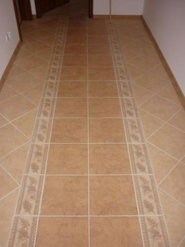 Beżowa podłoga z płytek ceramicznych ułożonych w prostokątnym wzorze z dekoracyjnym pasem pośrodku korytarza. Widok na całą długość podłogi.