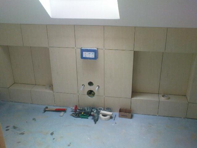 Łazienka w trakcie wykańczania: ułożone beżowe płytki ścienne, widoczne otwory na armaturę i narzędzia glazurnicze na podłodze. Skos sufitu z oknem dachowym.