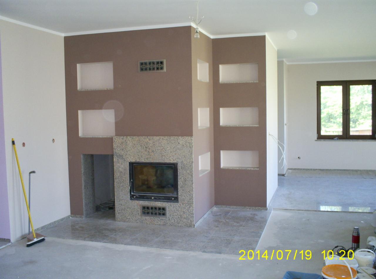 Wnętrze pokoju z kominkiem obudowanym kamieniem, wnęki w ścianie, ściany w odcieniach brązu i beżu, podłoga w trakcie prac wykończeniowych.