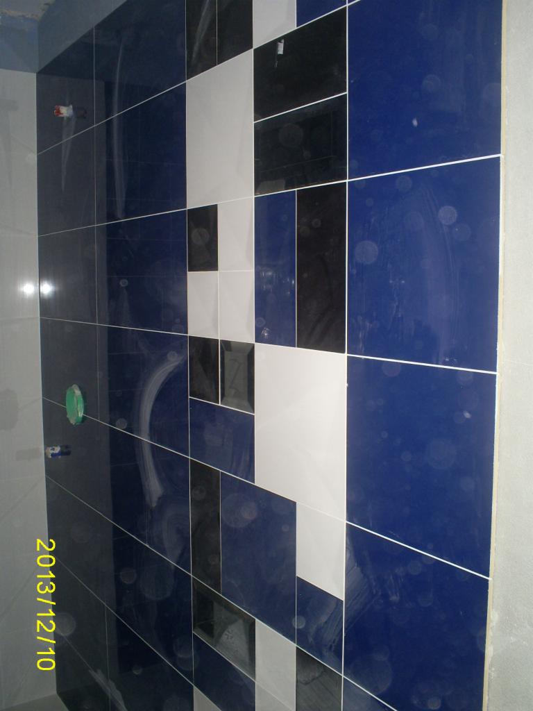 Ściana w łazience wyłożona błyszczącymi, kwadratowymi płytkami w odcieniach bieli, czerni i granatu, ułożonymi w geometryczny wzór. Widoczne fugi i fragment instalacji.