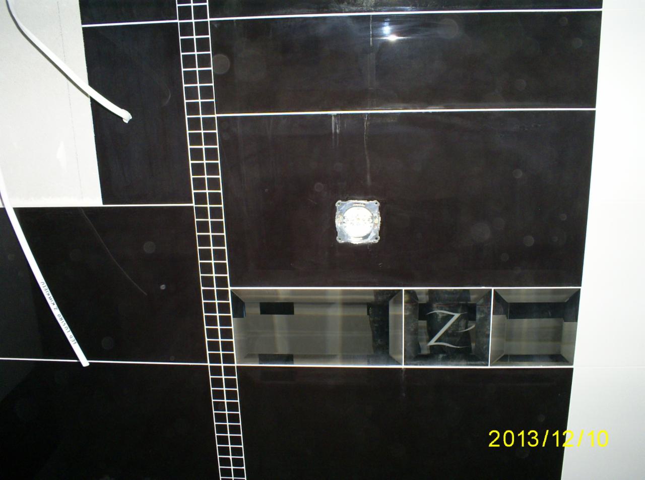 Czarne, błyszczące płytki z fugami i listwami, wnęka z literą 'Z', otwór na oświetlenie. Widoczne narzędzia glazurnicze. Ujęcie z dołu, perspektywa.