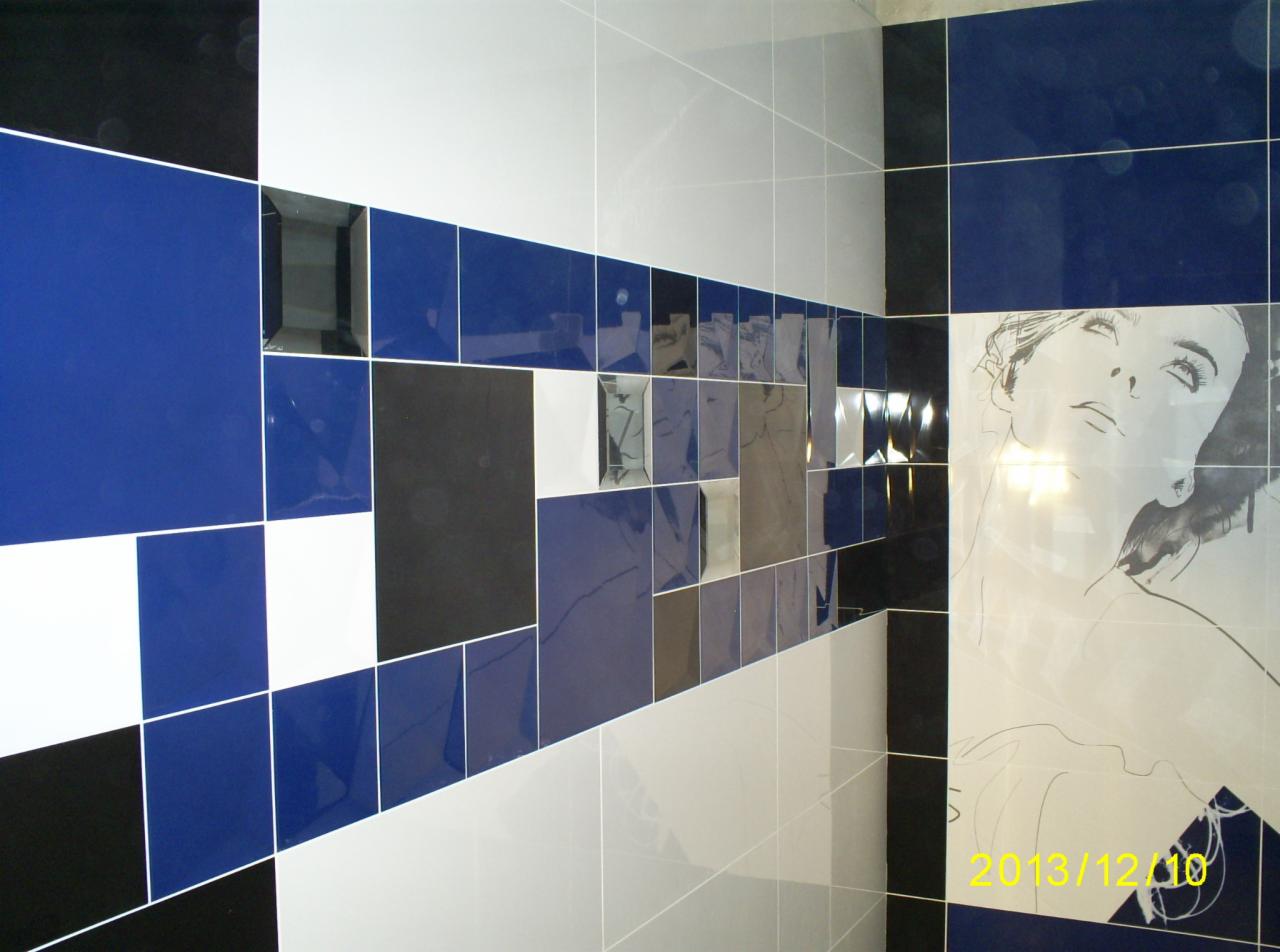 Ściana łazienki z mozaiką płytek w odcieniach bieli, szarości, czerni i kobaltu, z elementami lustrzanymi i grafiką kobiety. Ukończona realizacja.
