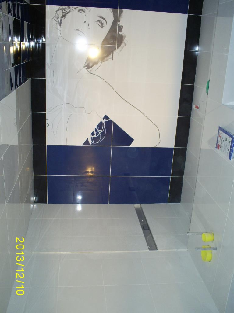 Wykończona łazienka z jasnymi płytkami i dekoracyjnym motywem kobiety na ścianie w odcieniach bieli i granatu. Widoczny odpływ liniowy w podłodze.