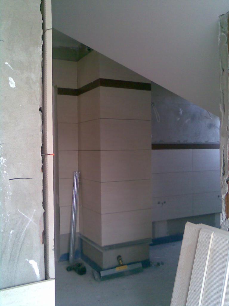 Wnętrze w trakcie prac wykończeniowych: obłożony płytkami filar z ciemnym paskiem, poziomica, zapas płytek. Widoczne niedokończone ściany.
