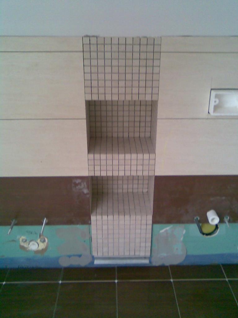 Ściana z beżowych płytek z wnęką wyłożoną mozaiką w łazience, na podłodze ciemne płytki. Widoczne elementy instalacji hydraulicznej przed wykończeniem.