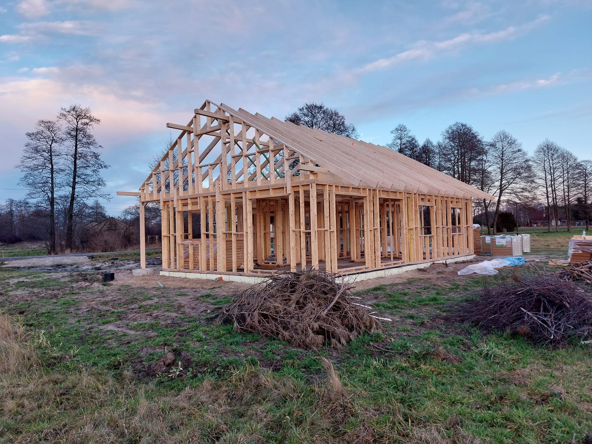 Konstrukcja drewniana domu w stanie surowym otwartym, widoczne elementy szkieletu, więźby dachowej, na zewnątrz stos gałęzi i materiały budowlane, pochmurne niebo w tle.