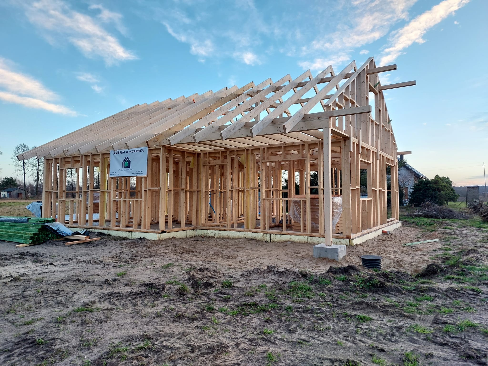Surowy stan domu w konstrukcji szkieletowej z widocznym ożebrowaniem dachu i logo firmy na ścianie, na tle błękitnego nieba z chmurami.