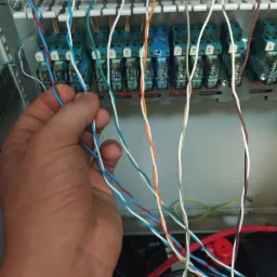 Ręka instalatora łączy kolorowe przewody w szafie sterowniczej z przekaźnikami, widoczne oznaczenia na przekaźnikach.