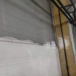 Częściowo wyczyszczona, pozioma, segmentowa brama przemysłowa w hali, z widocznym kontrastem między brudną g&oacute;rną częścią a czystą dolną. W tle widoczna konstrukcja dachu hali.