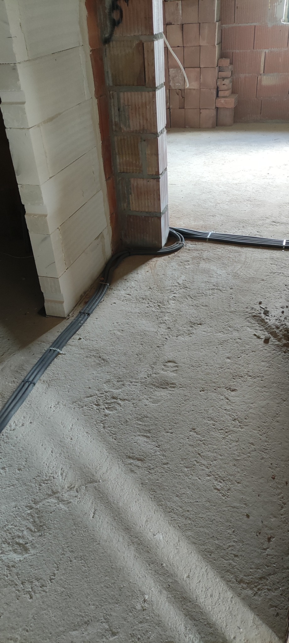 Szare rury w peszlach ułożone na surowym betonowym podłożu w nowym budynku, przymocowane opaskami zaciskowymi do podłoża i ściany z pustaków.