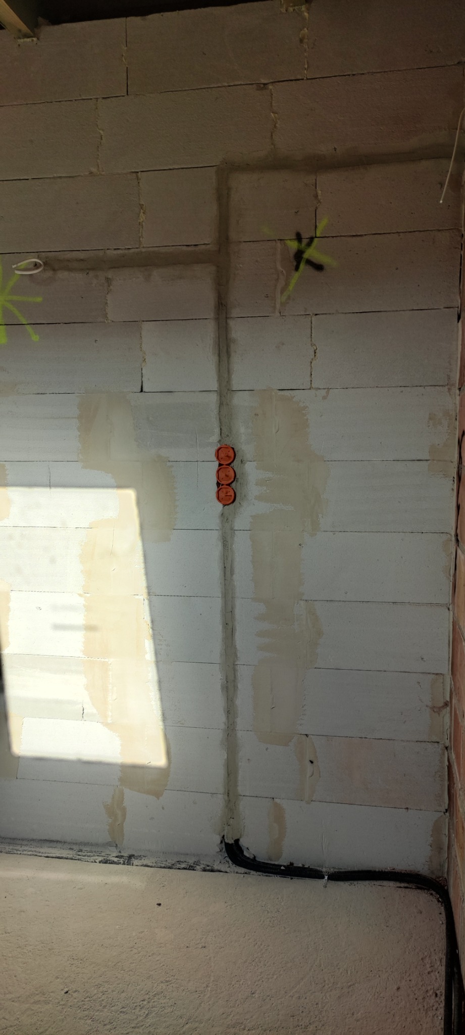 Ściana z betonu komórkowego z wyfrezowanymi bruzdami pod instalację elektryczną, widoczne pomarańczowe puszki pod gniazdka i peszel z kablami przy podłodze, oznaczona miejsca wierceń zielonym sprayem.