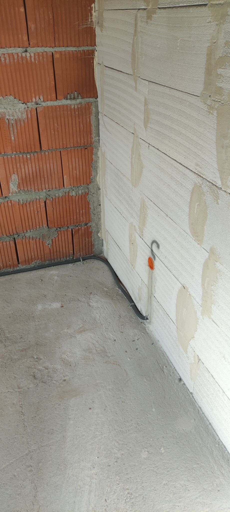 Fragment surowego wnętrza z widocznym narożnikiem, gdzie poprowadzono peszel elektryczny wzdłuż ściany z pustaków, zakończony pomarańczową puszką instalacyjną.