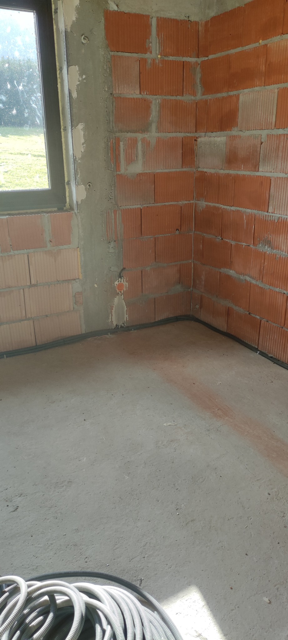 Wnętrze w trakcie budowy z pustaków ceramicznych, widoczne przewody elektryczne w peszlu ułożone wzdłuż ściany przy podłodze oraz puszka instalacyjna w ścianie.