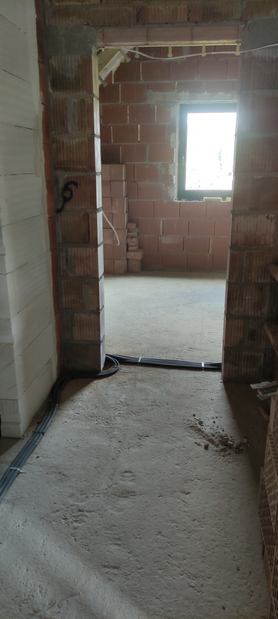 Surowy stan budowy: widok przez otwór drzwiowy na wnętrze z widoczną instalacją elektryczną w peszlach, ściany z czerwonej cegły i pustaków, betonowa podłoga w stanie surowym.