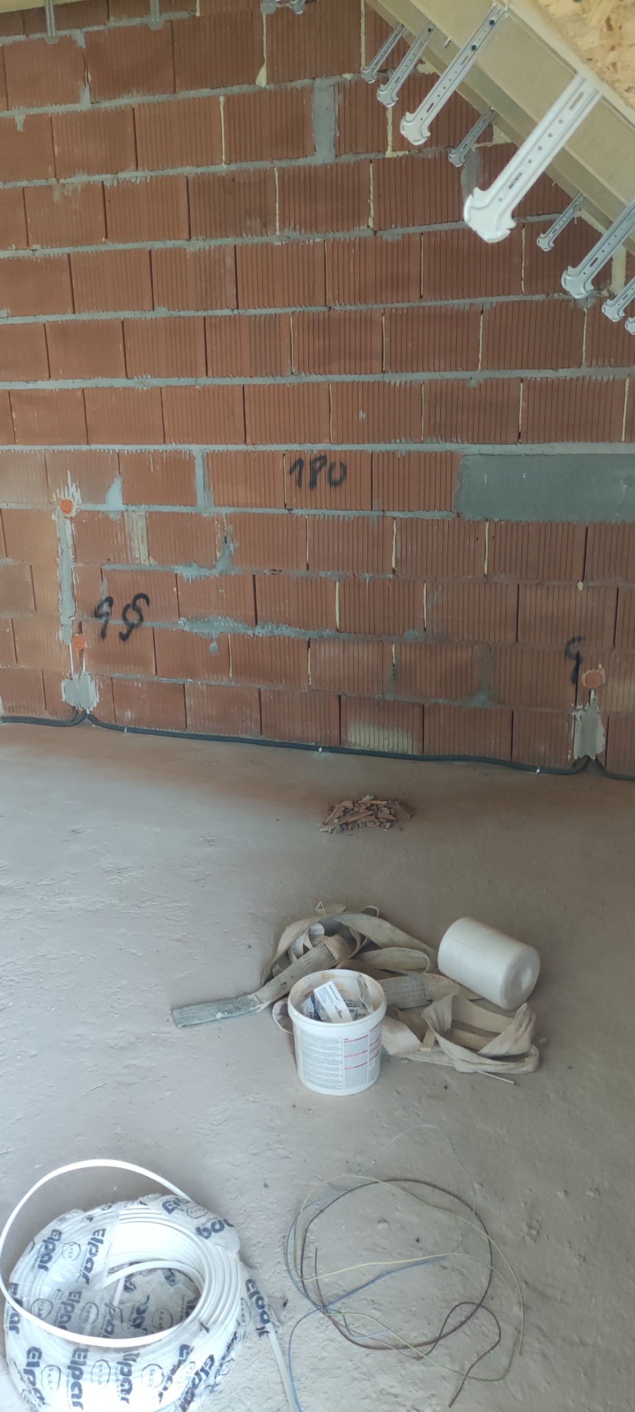 Wnętrze w trakcie prac instalacyjnych: widoczna ściana z czerwonej cegły z zaznaczonymi punktami elektrycznymi, przewody elektryczne prowadzone wzdłuż ściany, na podłodze leżą zwinięte kable...