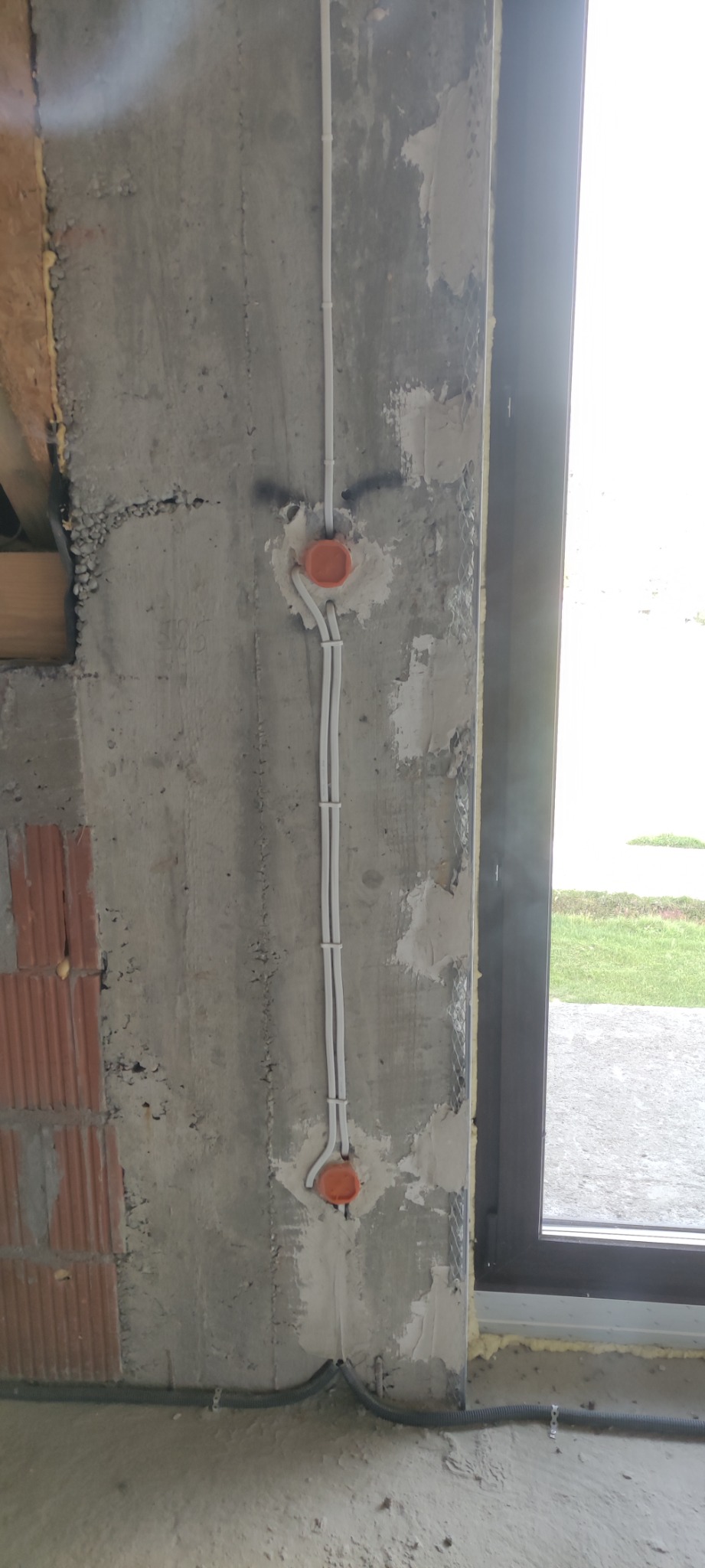 Surowy betonowy mur z zamontowanymi puszkami elektrycznymi i przewodami w białej izolacji, widoczne elementy muru z cegły i okno.