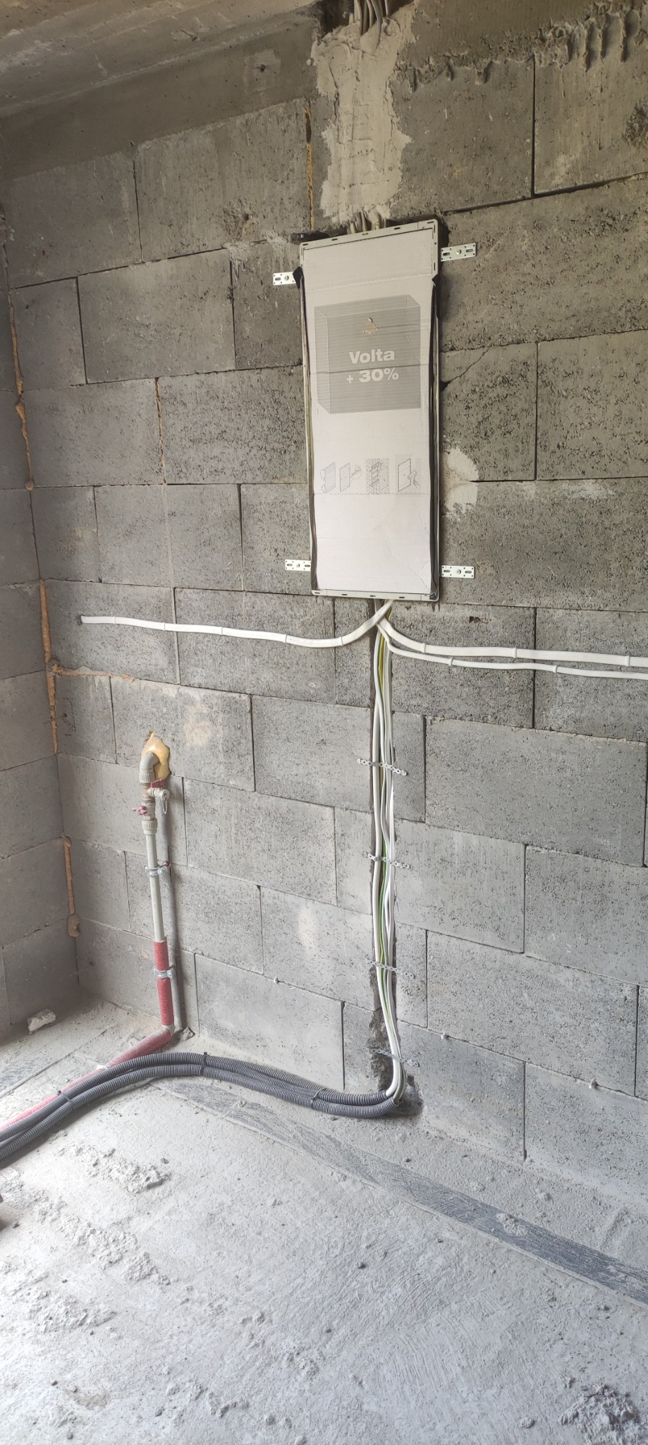 Surowa ściana z betonu komórkowego z zamontowaną skrzynką elektryczną Volta +30% i poprowadzonymi kablami elektrycznymi, widoczne także rury instalacji wodnej w rogu pomieszczenia.