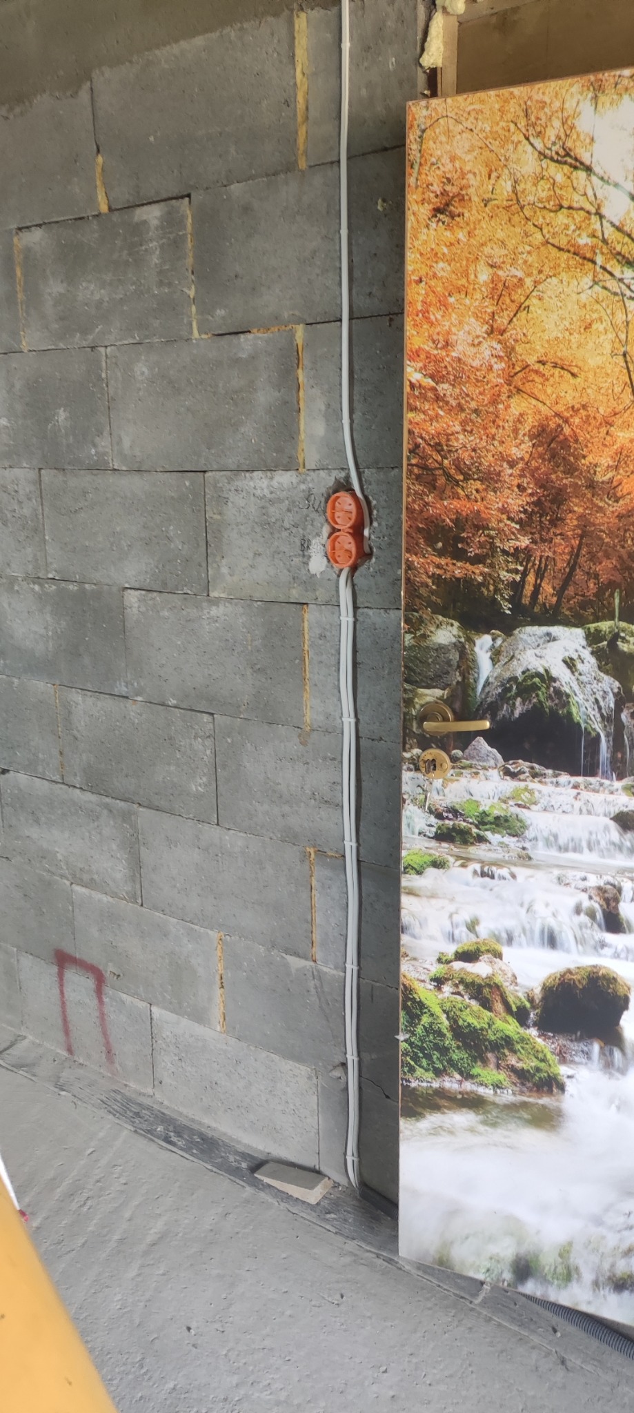 Szara ściana z betonu komórkowego z pomarańczowymi puszkami elektrycznymi i białymi przewodami, obok drzwi z fototapetą przedstawiającą jesienny las z wodospadem.