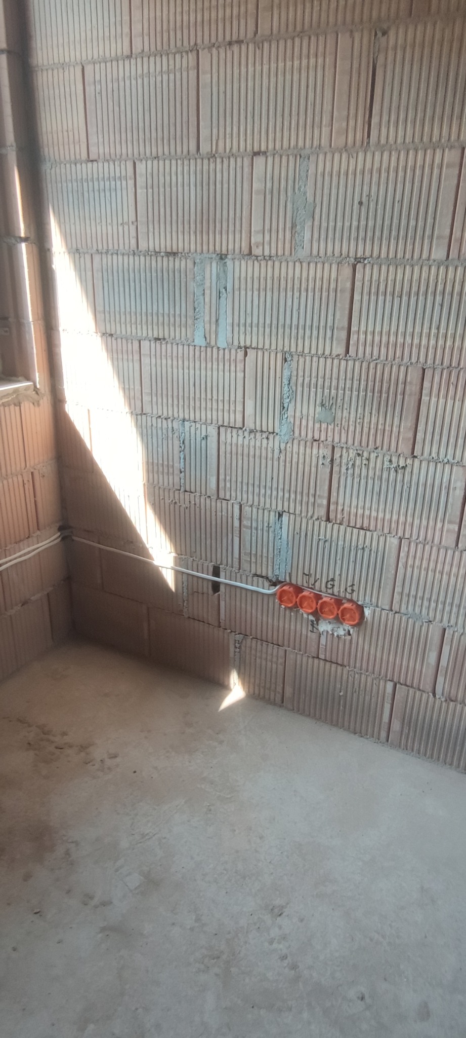 Wnętrze w trakcie budowy z widocznymi przewodami elektrycznymi w rurce i puszkami pod gniazdka, ściany z czerwonej cegły dziurawki, surowa podłoga.