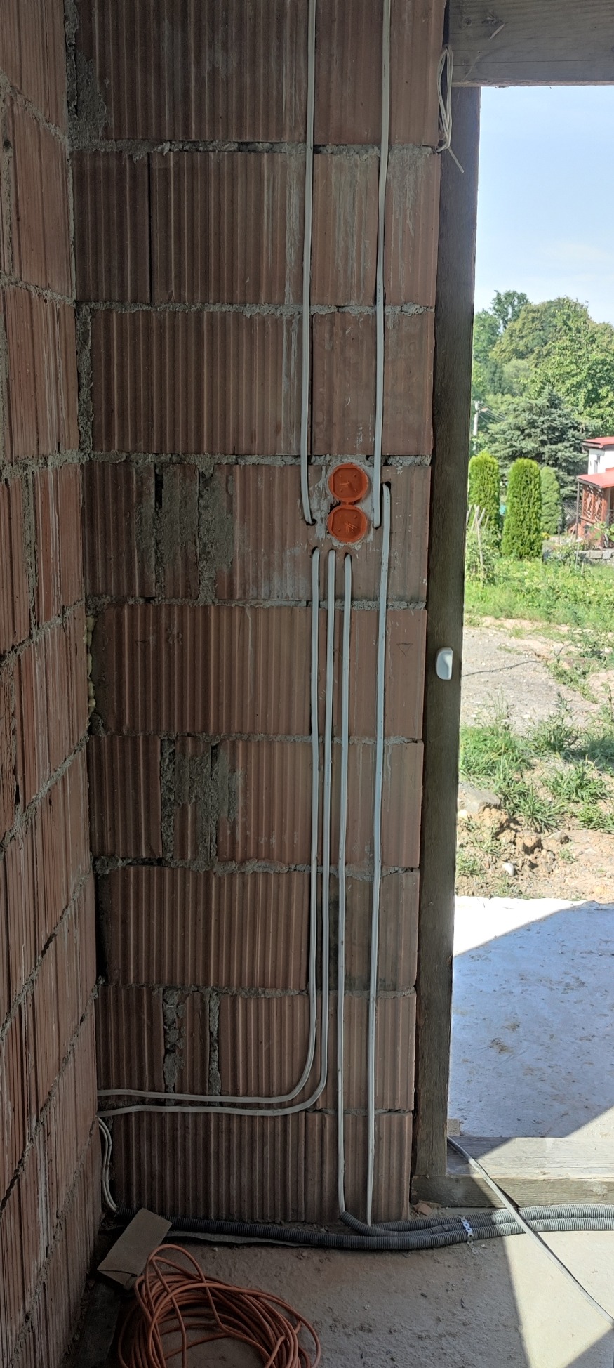 Wewnętrzna instalacja elektryczna w trakcie montażu, widoczne przewody prowadzone wzdłuż ściany z pustaków ceramicznych, pomarańczowe puszki pod gniazdka i zwój kabla na podłodze.
