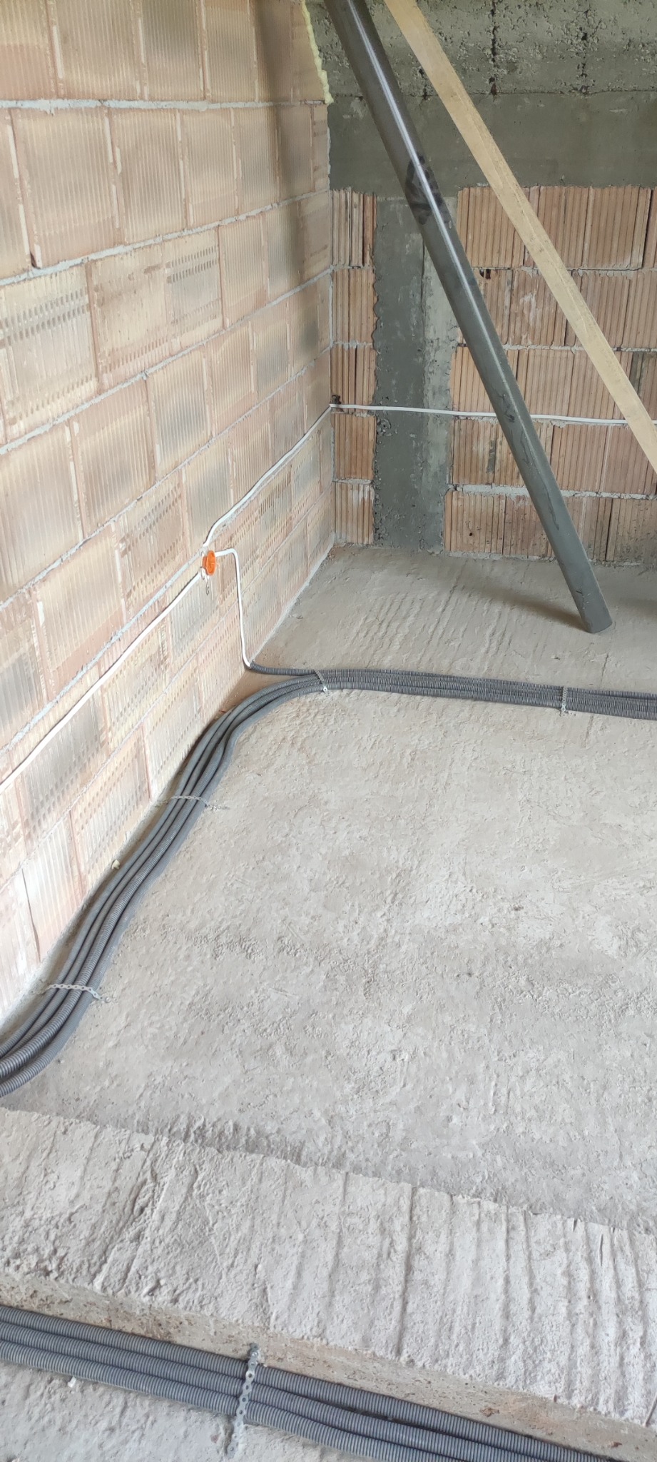 Surowy stan: Kable elektryczne w szarych peszlach, mocowane metalowymi uchwytami do surowej ściany z cegły dziurawki i wylewki betonowej, widoczna puszka instalacyjna. Rury instalacyjne w tle.