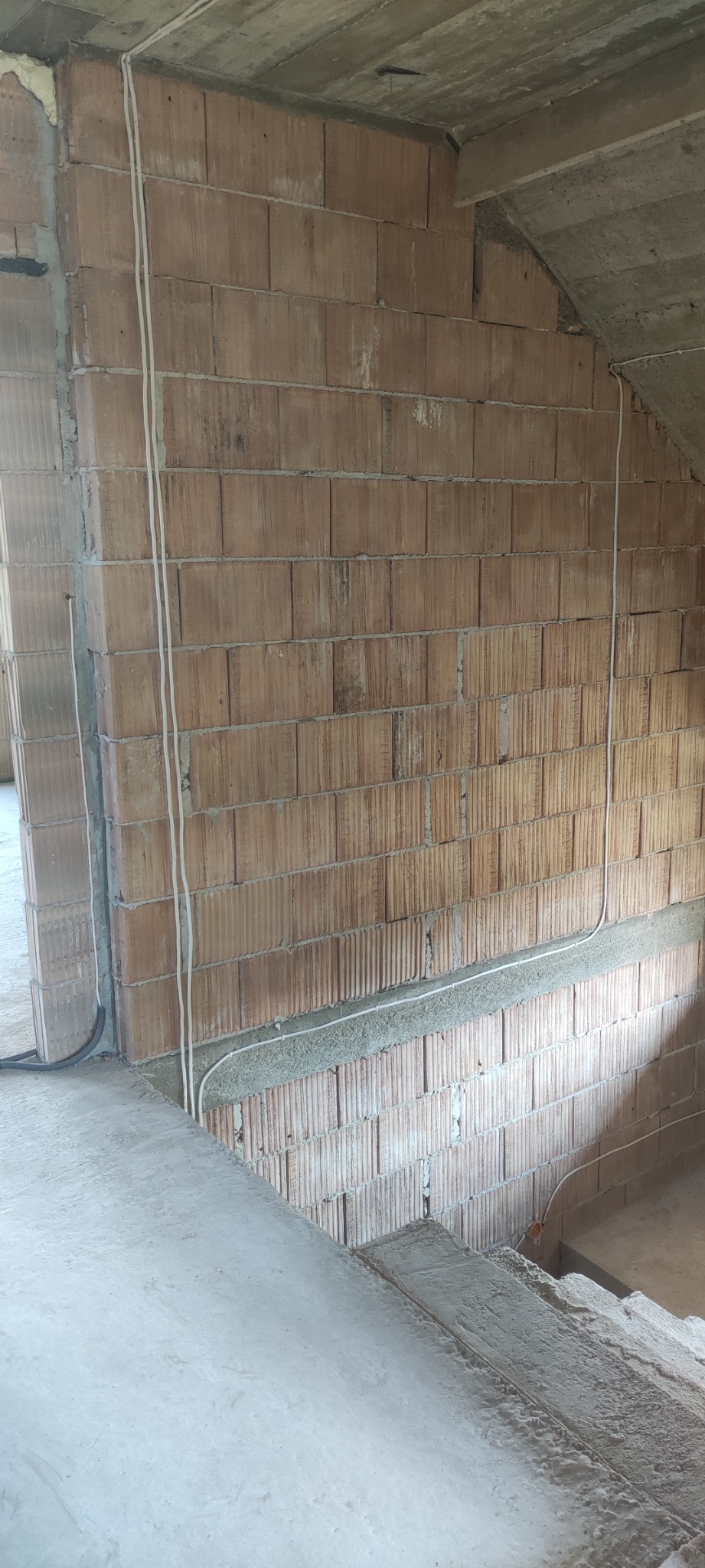 Surowe wnętrze w trakcie budowy, widoczne przewody elektryczne poprowadzone po ścianie z cegieł dziurawki, betonowy strop i schody.