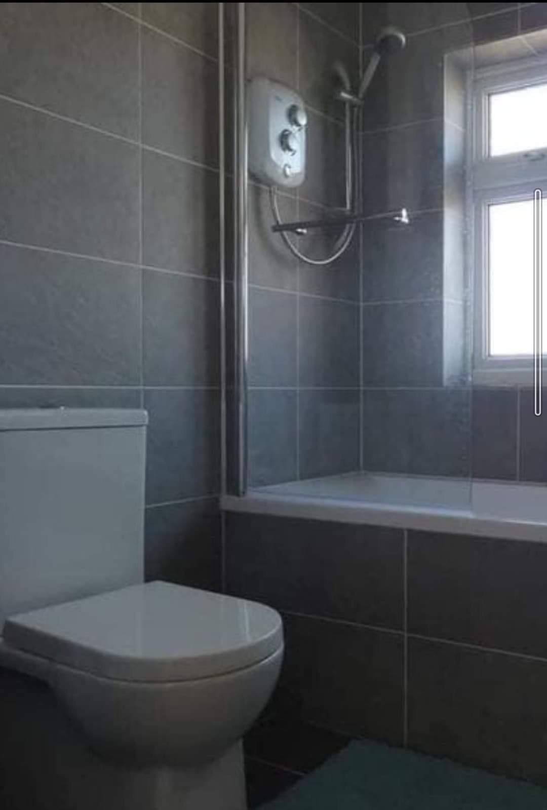 Szara łazienka z wanną, prysznicem i toaletą, wykończona płytkami w odcieniach szarości, z oknem w tle.