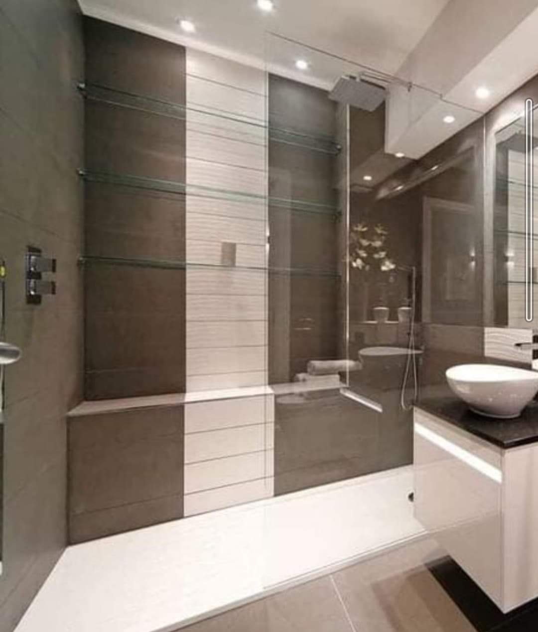 Nowoczesna łazienka z kabiną prysznicową typu walk-in, wykończona płytkami w odcieniach szarości i bieli, z szklanymi półkami i umywalką nablatową.