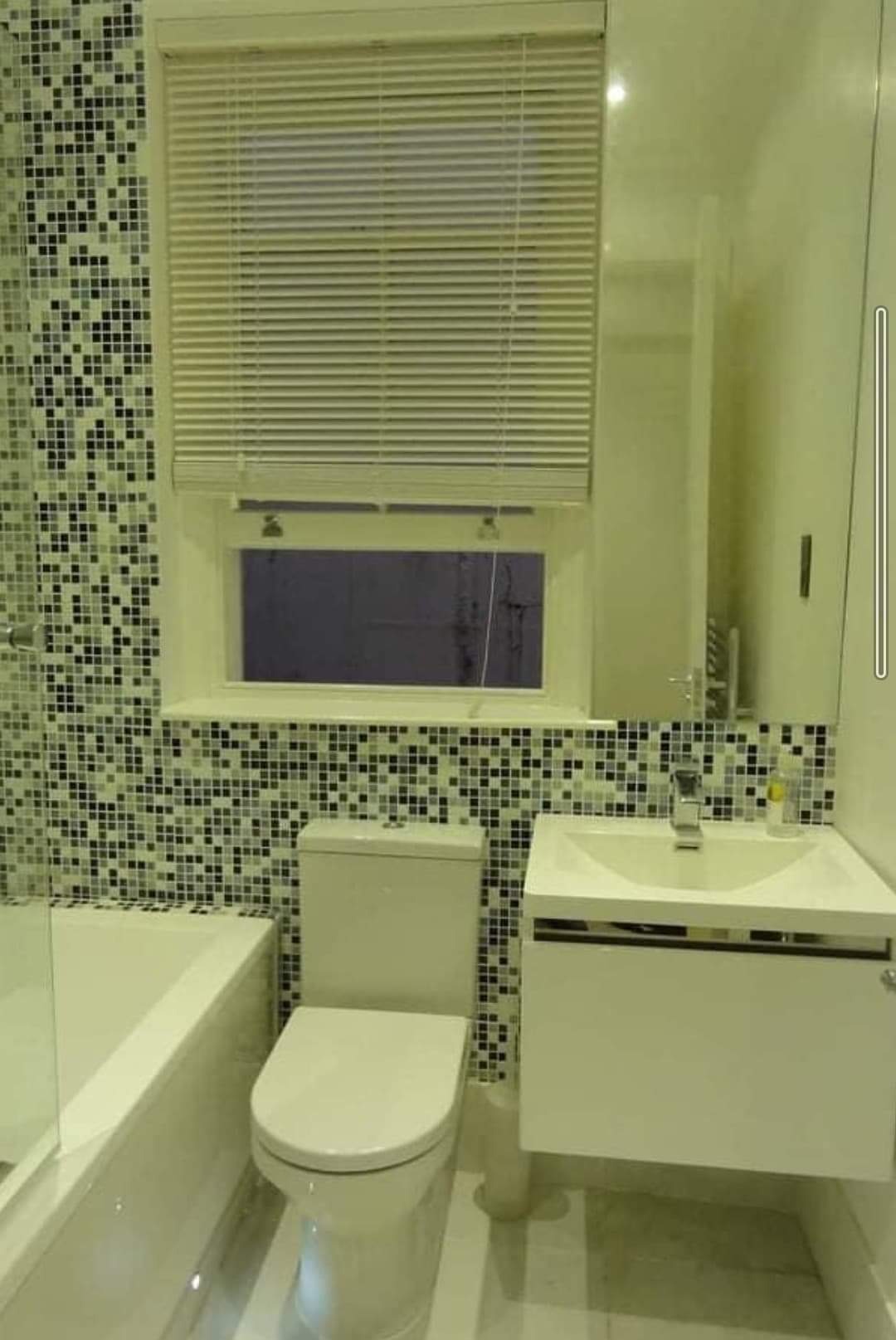 Nowoczesna łazienka z mozaikową ścianą, białą armaturą i roletami w oknie.