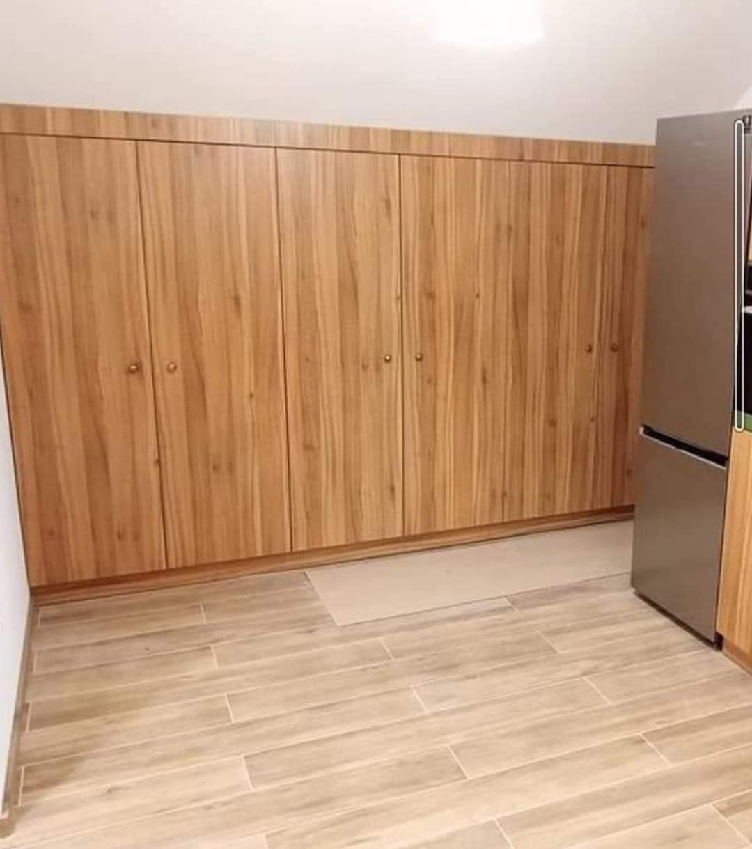 Zabudowa kuchenna z drewnianymi frontami, dopasowana do skosu dachu, obok stoi lodówka w kolorze stali nierdzewnej, na podłodze płytki imitujące drewno.