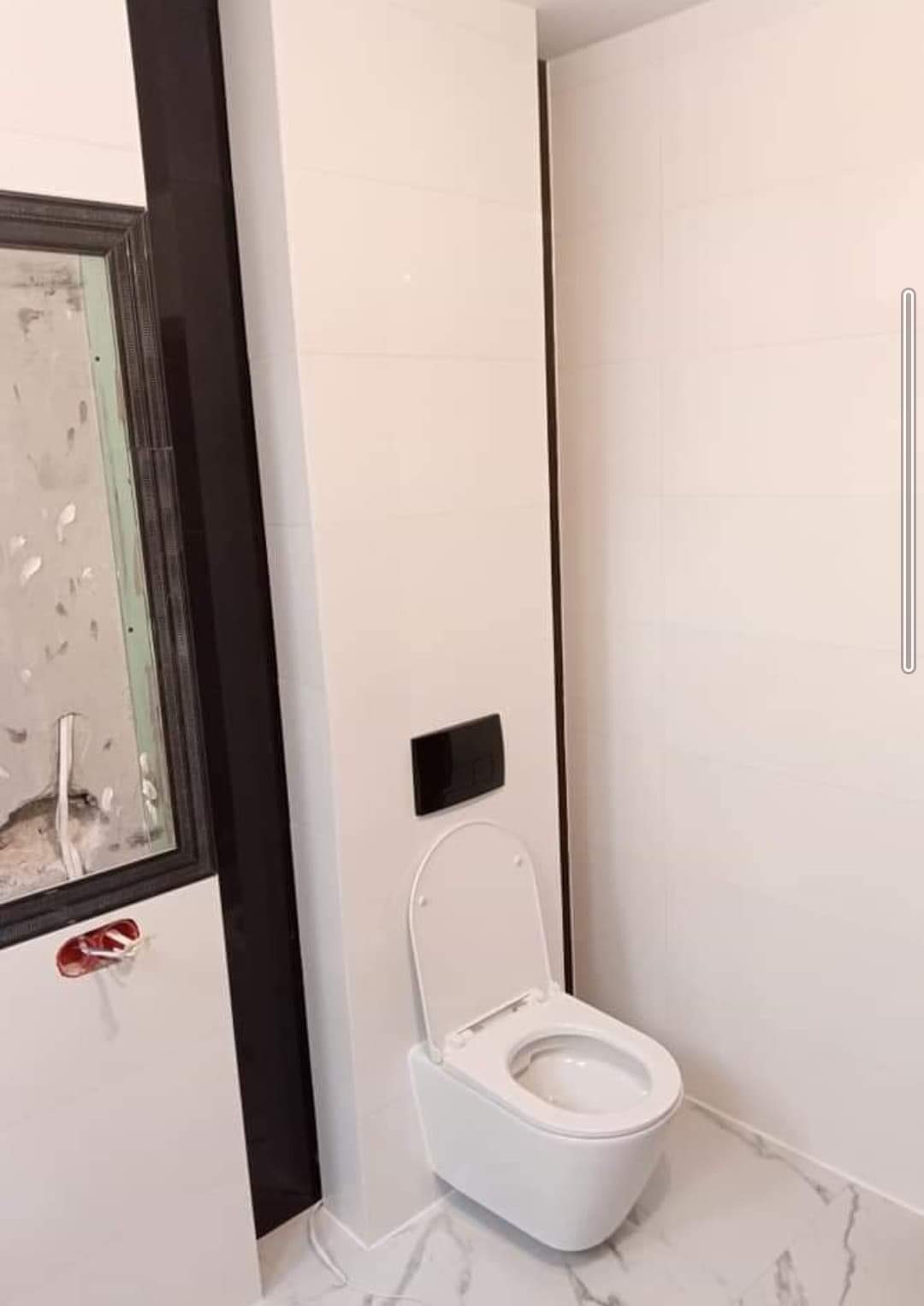 Nowoczesna, podwieszana toaleta z czarnym przyciskiem spłukującym na tle białych płytek, z fragmentem lustra w trakcie montażu po lewej stronie, na podłodze marmuropodobne płytki.