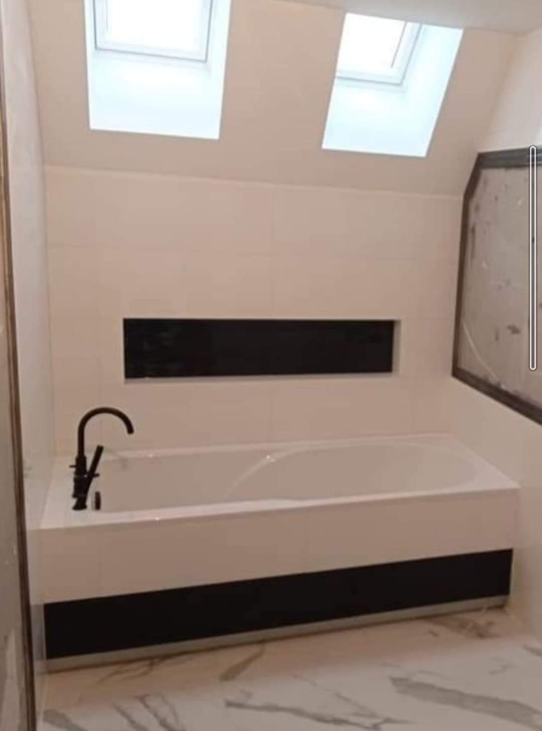 Nowoczesna łazienka na poddaszu z białą wanną obudowaną czarnymi panelami, czarną armaturą i wnęką w ścianie wyłożoną czarnymi płytkami, oświetlona przez okna dachowe.