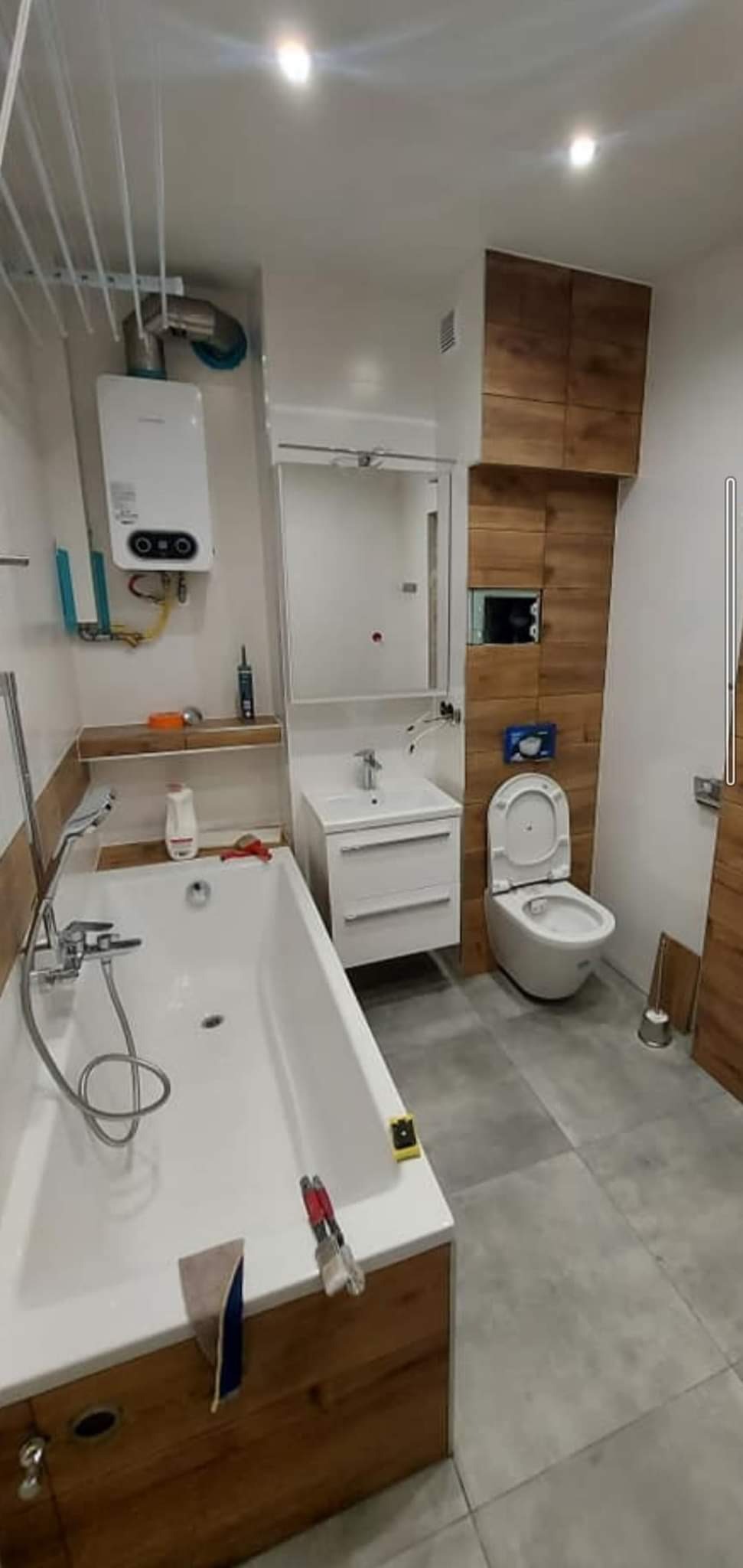 Wykończona łazienka z wanną, umywalką z szafką, toaletą i bojlerem. Ściana wykończona płytkami imitującymi drewno.