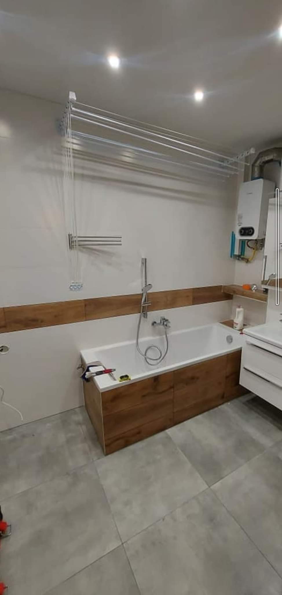 Wykończona łazienka z wanną obudowaną panelami imitującymi drewno, szarą podłogą z płytek, grzejnikiem i suszarką podsufitową. Na wannie leżą narzędzia.