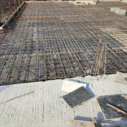 Zbrojenie stropu domu jednorodzinnego, widok z góry na siatkę z prętów stalowych na szalunku, w tle zabudowa jednorodzinna i krajobraz.