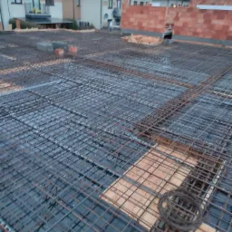 Zbrojenie stropu z siatki stalowej na budowie domu jednorodzinnego, widoczne bloczki z czerwonej cegły silikatowej w tle, pochmurne niebo.