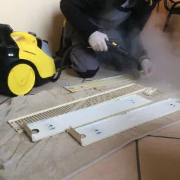 Czyszczenie parowe elementów grzejnika przez osobę w rękawicach ochronnych, na podłodze leży beżowy ręcznik, obok żółto-czarny odkurzacz parowy.